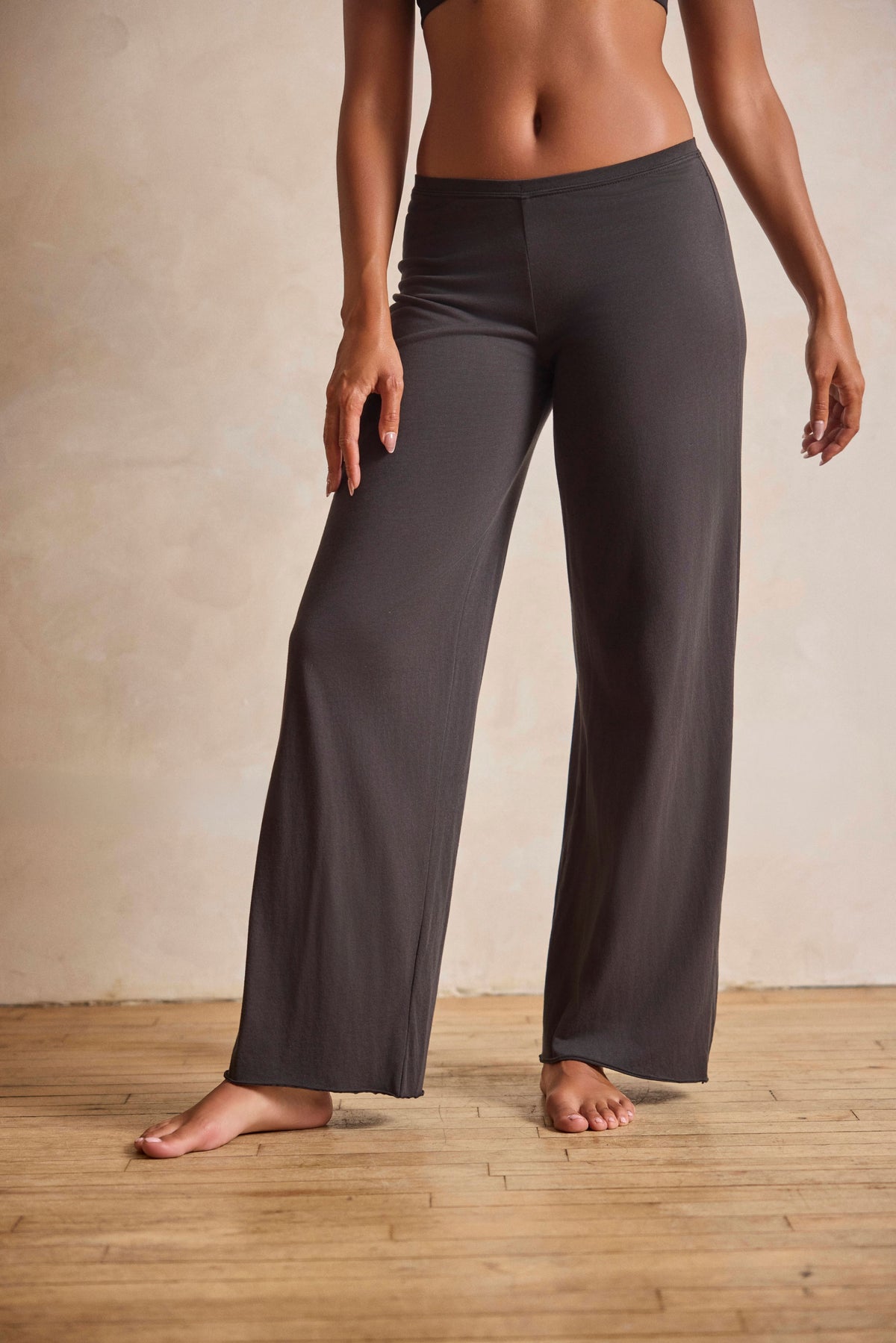 A model wearing dark charcoal gray wide-leg yoga pants, standing barefoot on a light wood floor against a textured beige wall.