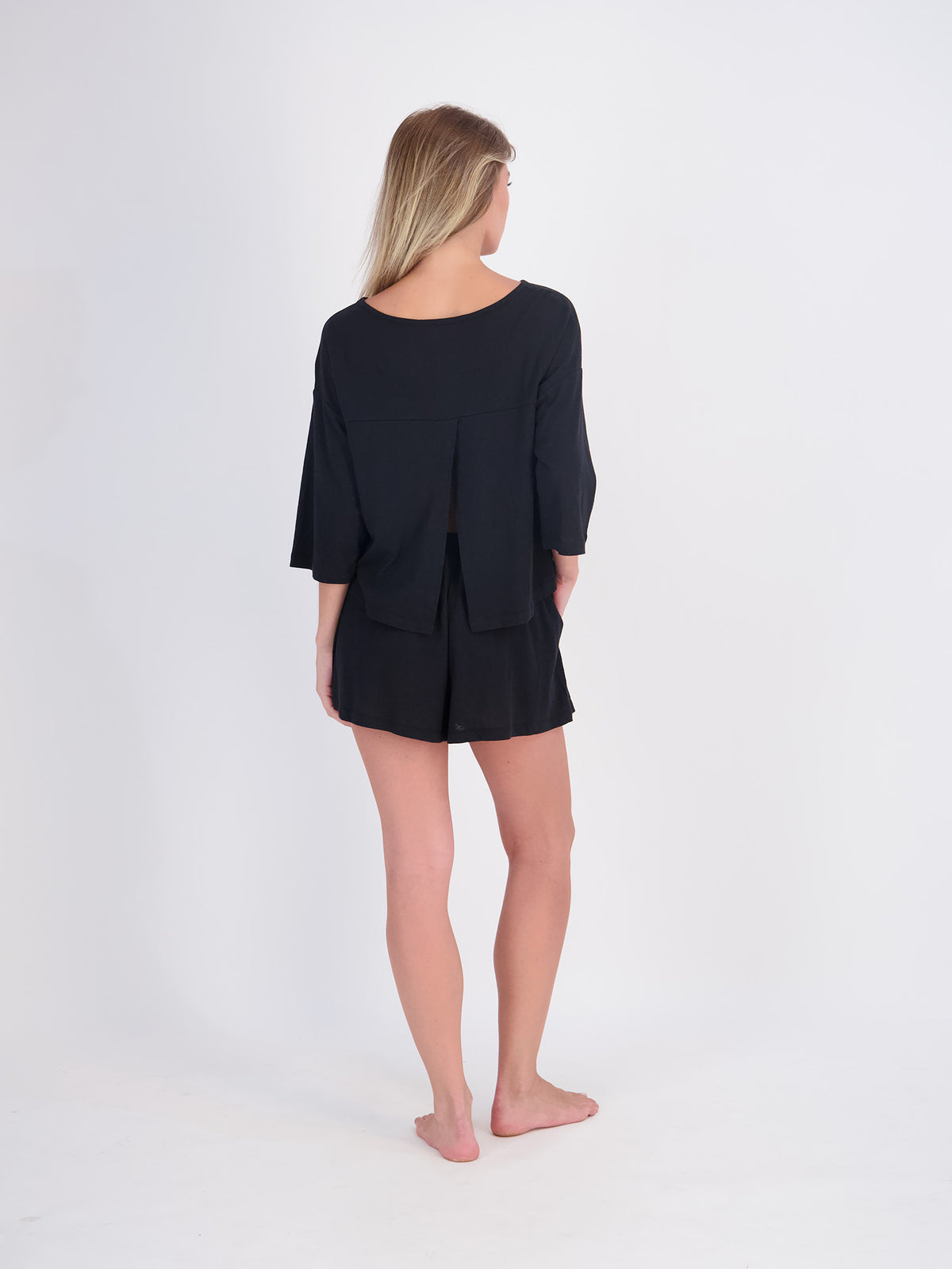 A woman with blonde hair is shown from the back wearing a black three-quarter sleeve top and matching black shorts, standing barefoot against a plain white studio background.