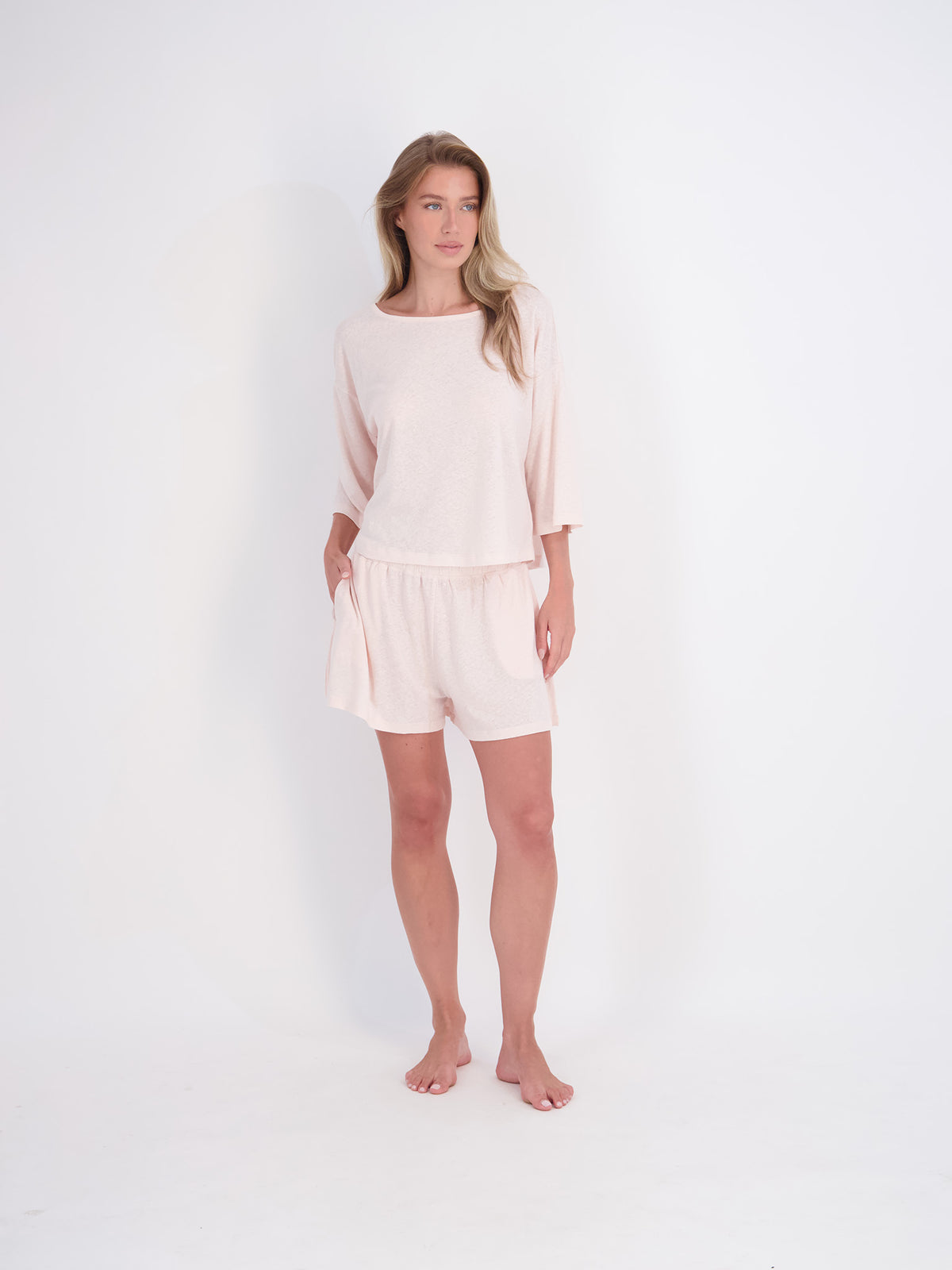 A woman modeling a light pink two-piece lounge set consisting of a loose three-quarter sleeve top and matching shorts, standing against a seamless white studio background.