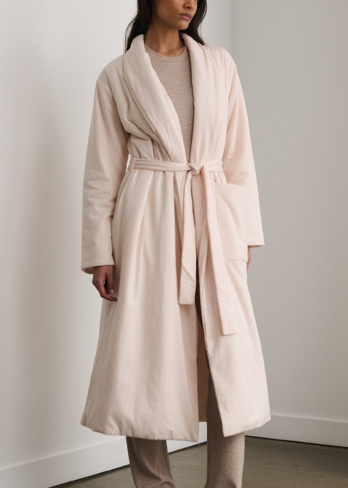 A woman standing outdoors wearing a white robe with a belt and three pockets, with a plant in the background.